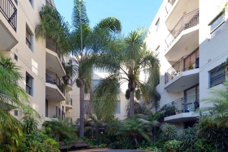 A tranquil courtyard is surrounded by residential buildings, featuring tall palm trees and lush greenery. The well-maintained space offers a glimpse of the serene atmosphere, with the pool area partially visible in the center, inviting relaxation amid nature.