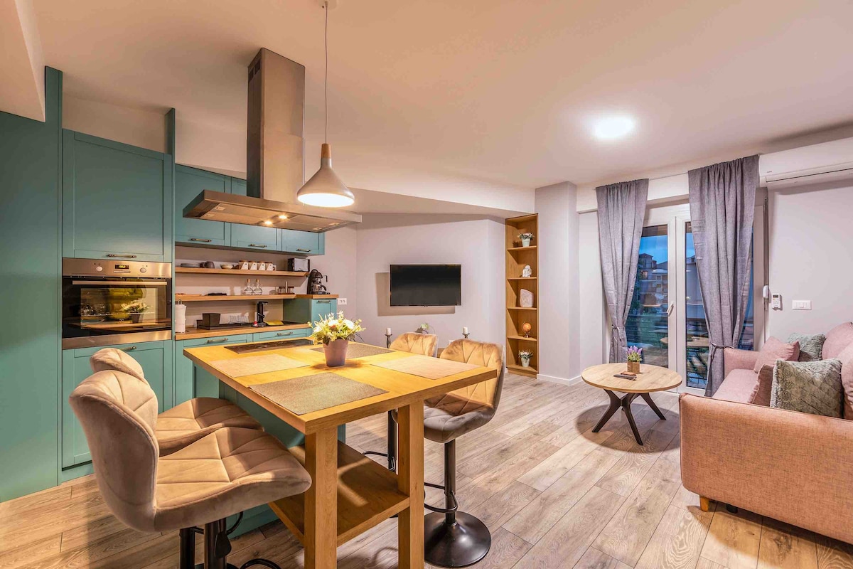 A modern kitchen area features sleek cabinetry and a spacious dining table with four upholstered chairs. A well-equipped cooking space includes an oven and microwave, while a cozy seating area is highlighted by a light-colored sofa and decorative pillows. Natural light filters through sheer curtains.