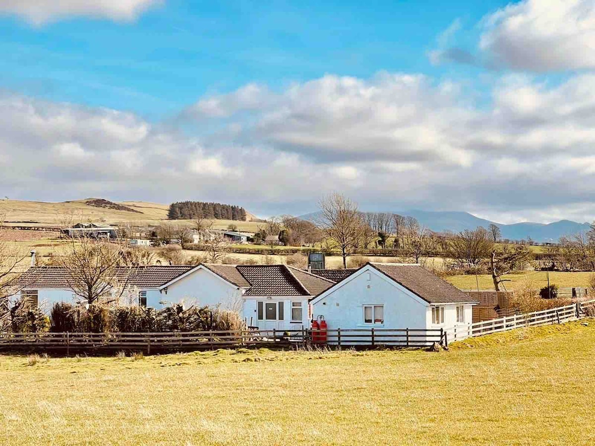 A view of a quaint holiday apartment highlighted by its white exterior and gently sloping roof. The surrounding landscape features open fields with distant hills and a cloudy sky, creating a serene setting. A wooden fence complements the natural scenery.