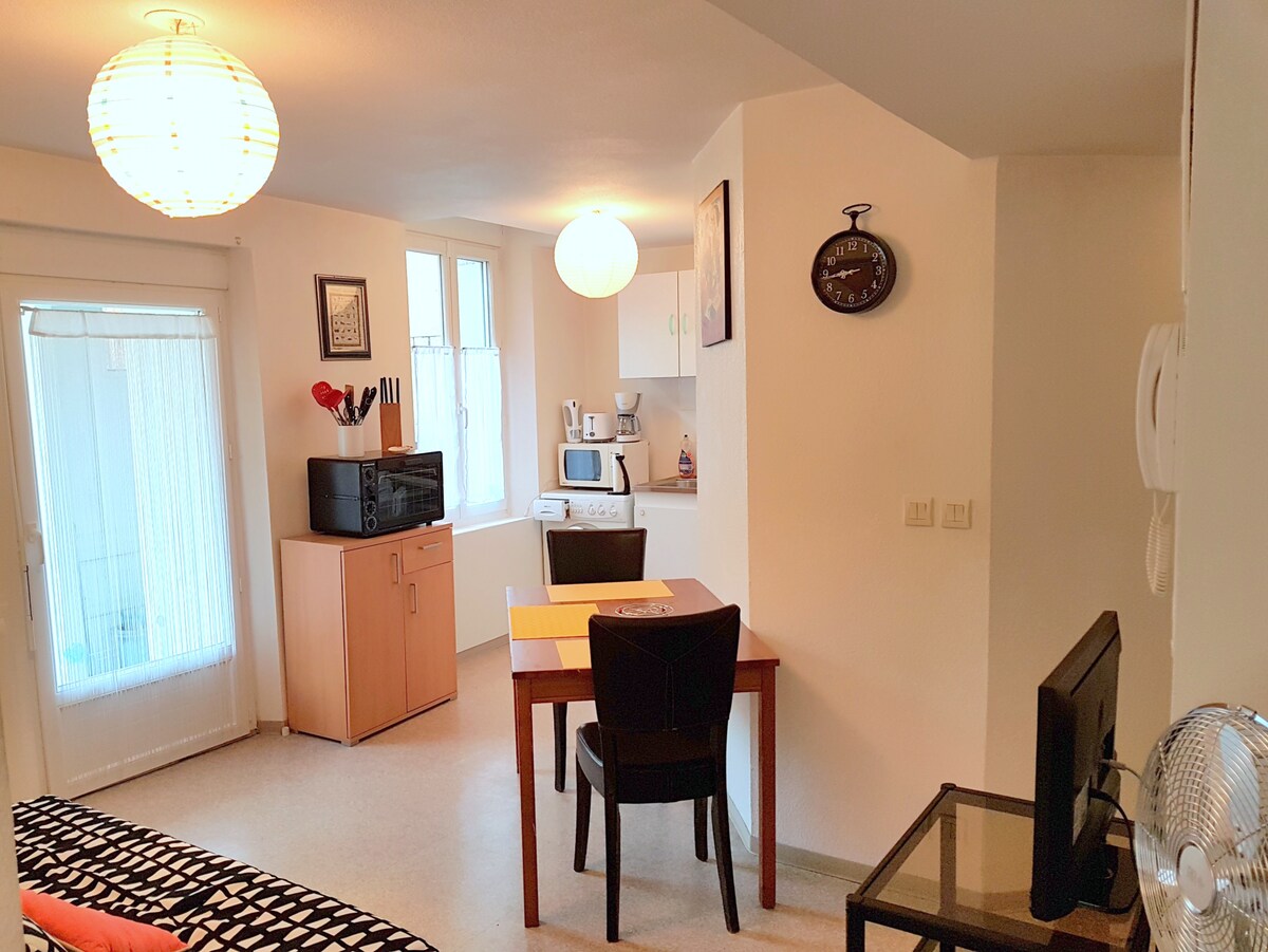 A well-lit living area features a small dining table set for two, complemented by a black chair. In the background, a kitchenette with essential appliances is visible, alongside a microwave and a black cabinet. Light streams through a glass door leading to the balcony.