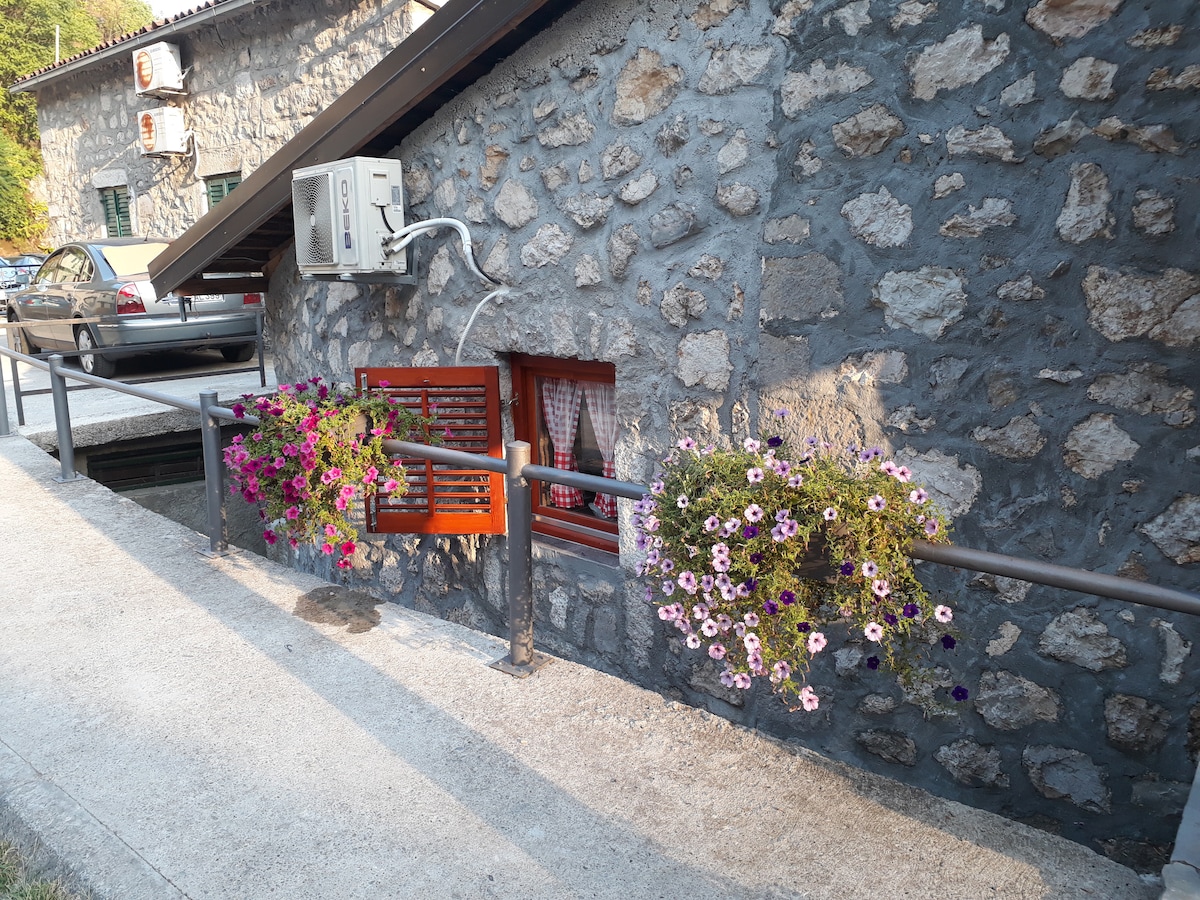 A stone wall features a wooden window with red shutters, partially opened to reveal a curtain. Colorful flower boxes filled with blooming petunias and greenery adorn a railing along a paved walkway, providing a charming touch to the entrance.