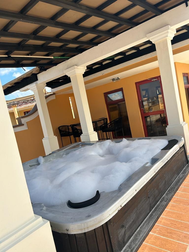 A private hot tub is featured on the outdoor terrace, surrounded by a pergola supported by white columns. Bubbles cover the water surface, while a comfortable seating area with dark chairs is set nearby. The warm yellow walls enhance the inviting atmosphere.