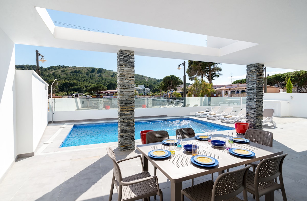 An outdoor terrace features a dining table set for six, complemented by a scenic view of the private pool and surrounding greenery. The poolside area includes sun loungers, while natural light filters through the overhead skylight, enhancing the space's openness.