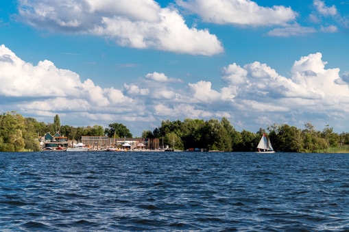 A serene view of a body of water surrounded by lush greenery, showcasing a few sailboats gently navigating the waves. In the background, a collection of buildings can be seen, reflecting the natural beauty of the setting under a blue sky dotted with white clouds.