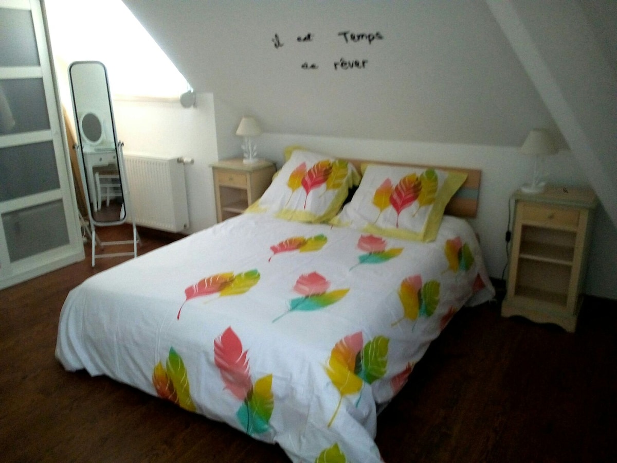 The bedroom features a spacious bed adorned with a floral patterned duvet cover. Light-colored bedside tables are positioned on either side, each topped with a lamp. A full-length mirror reflects natural light, and a wall displays the phrase 'Il est temps de rêver' in elegant script.