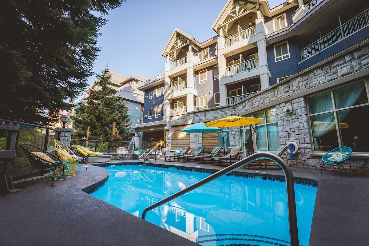 A clear outdoor pool is framed by a serene landscape, with comfortable lounge chairs placed around its perimeter. The multi-story building in the background features balconies and is surrounded by lush trees, creating a tranquil setting for relaxation.