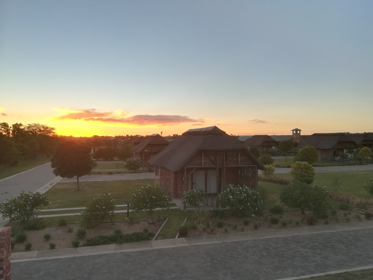 The image captures a tranquil sunset over a landscape of chalets with thatched roofs. Lush gardens and well-maintained pathways are visible, leading to the accommodations. The sky is painted with warm hues of orange and pink, creating a serene backdrop for the setting.