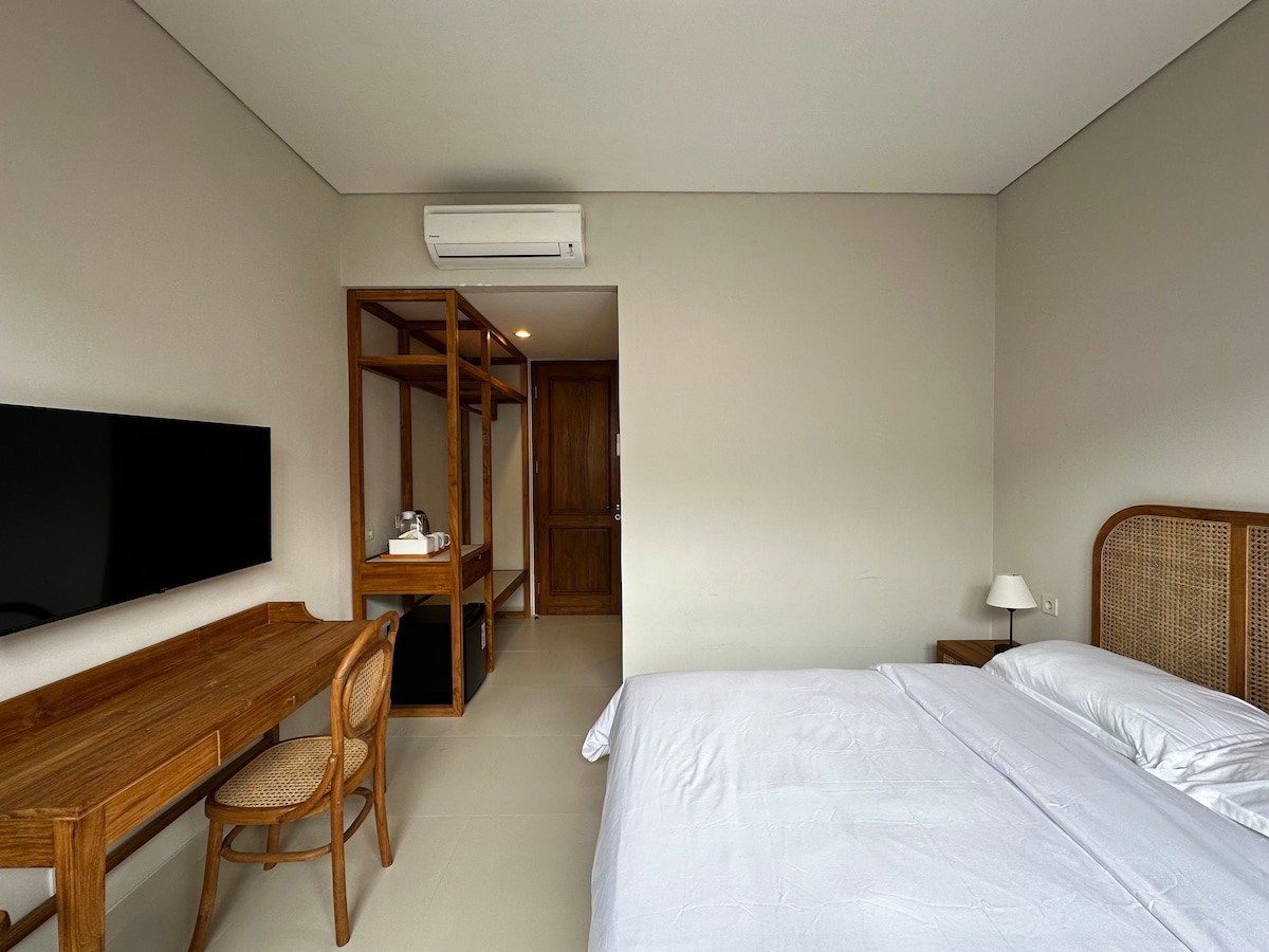 A neatly arranged room features a spacious bed with crisp linens, complemented by a wooden desk and a comfortable chair. A wall-mounted television is positioned opposite the bed. Open shelving and a closet are visible, with a doorway leading outside.