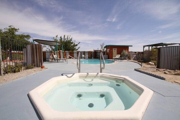 A hot tub with steps leads to a clear, inviting pool area, surrounded by a stone and gravel landscape. Sun loungers are positioned nearby, offering a relaxing space. The backdrop features an open sky and lush desert vegetation.