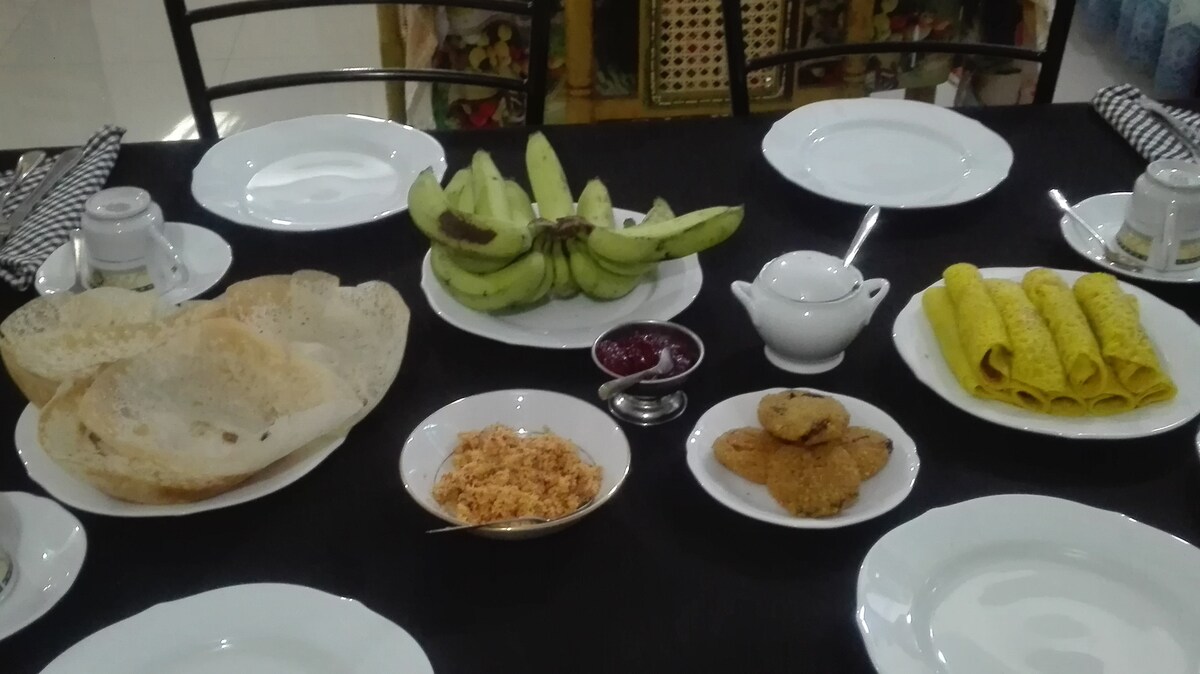 A dining table is set with several plates featuring a variety of local dishes. A bowl of rice flour pancakes and a platter of bananas are visible, along with a serving of chutney and crispy snacks. The table is dressed with a dark tablecloth and white dinnerware.