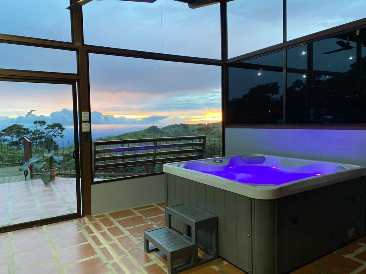 A hot tub with adjustable lighting is positioned beside large glass windows, offering views of a vibrant sunset. The floor is tiled in warm tones, and wooden seating is present near the entrance. The serene outdoor landscape features trees and distant hills.