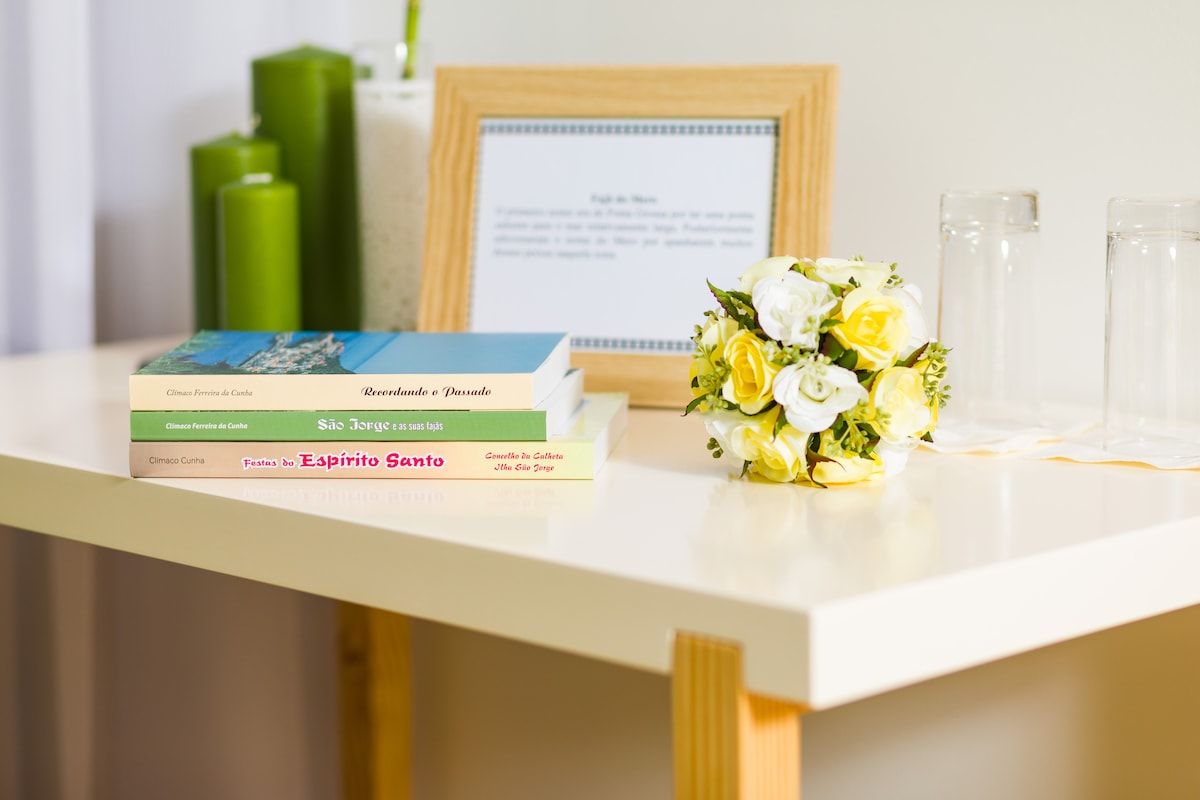 A small table is arranged with three neatly stacked books featuring colorful covers. A decorative flower bouquet of yellow and white blooms is placed beside the books. Green candles and a framed informational card are positioned in the background, adding to the understated decor.