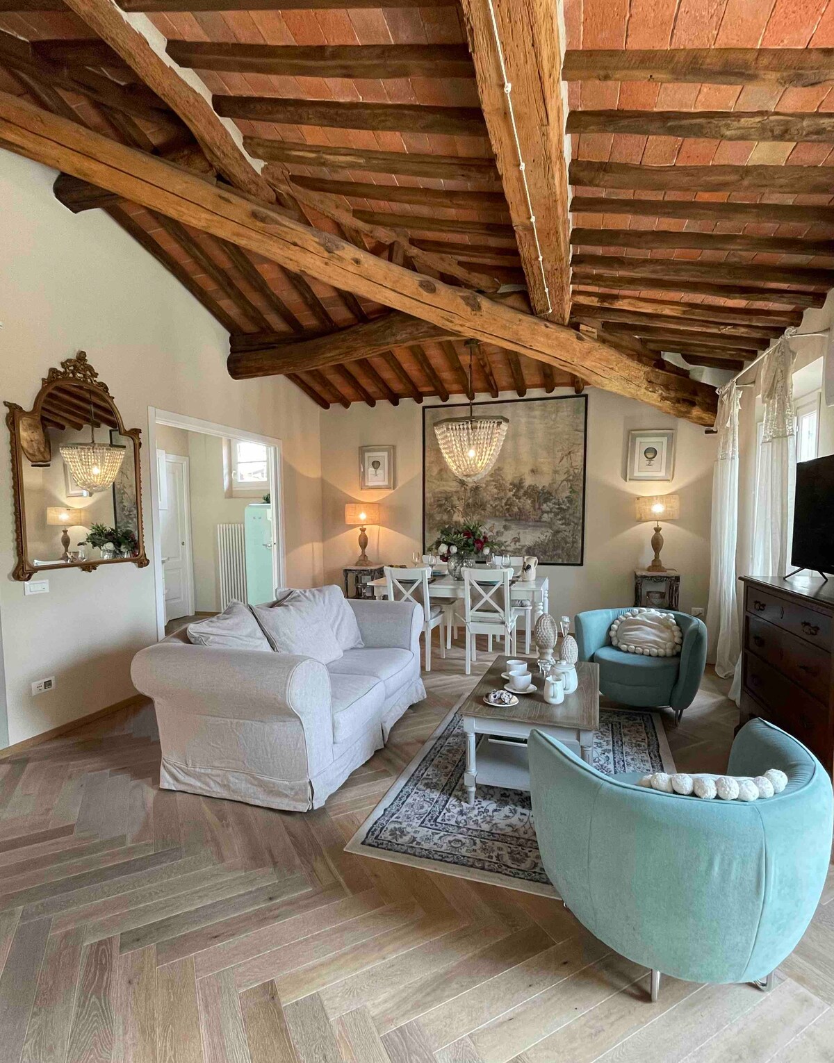 A spacious living area features a combination of light-colored seating and elegant furnishings. Natural light enters through large windows, illuminating the wooden beams above. A dining area is visible in the background, complemented by a decorative wall art piece and soft ambient lighting.