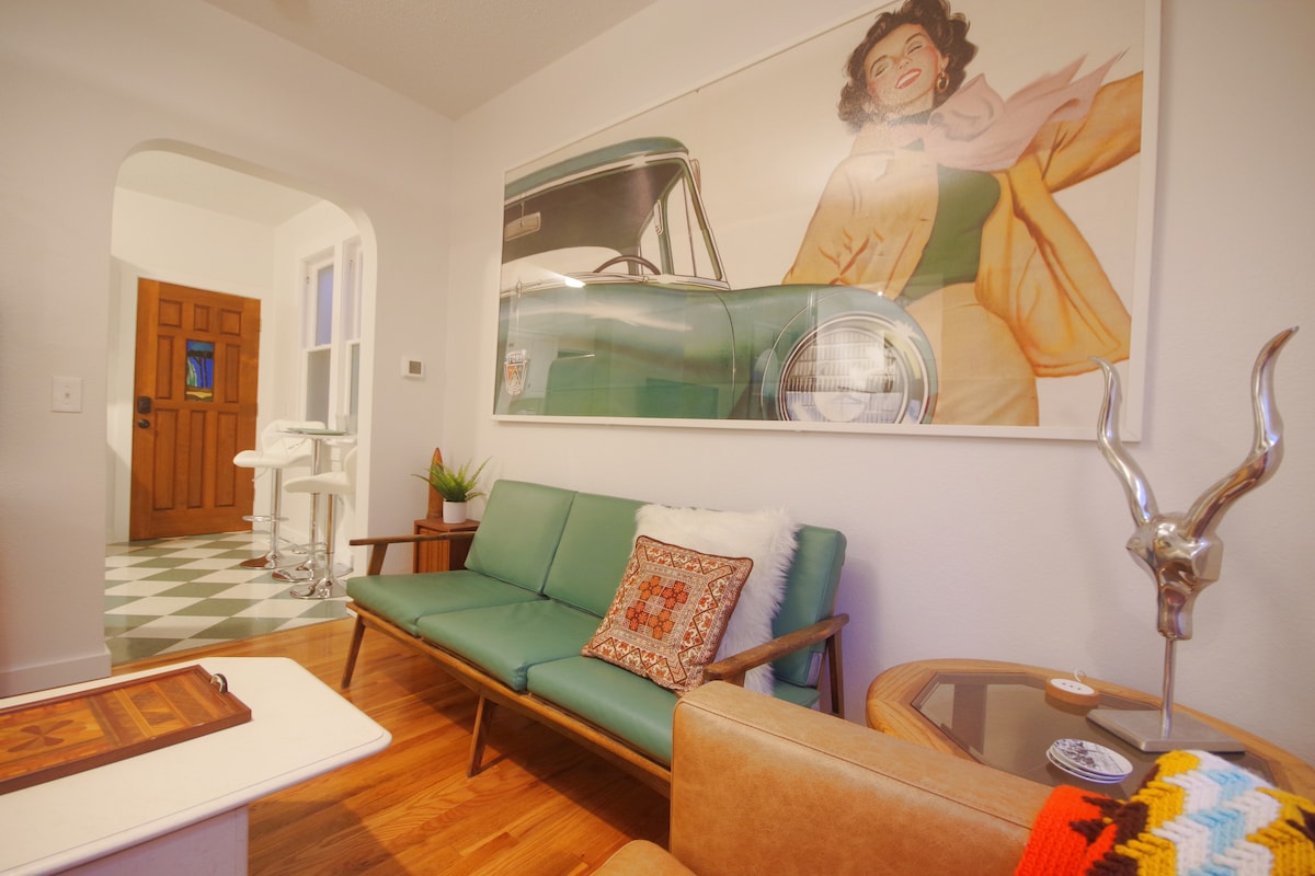 A living area is highlighted by a vintage green couch with wooden legs. A decorative pillow adds a touch of comfort. A large artwork featuring a classic car adorns the wall, while a doorway leads to an additional space visible in the background.