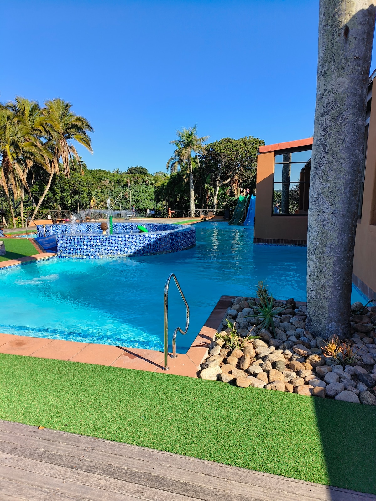 A vibrant pool area is surrounded by lush greenery, featuring a water slide, multiple zones for relaxation, and a well-maintained garden element with decorative rocks. Sunlight sparkles on the turquoise water, creating a refreshing atmosphere for guests.
