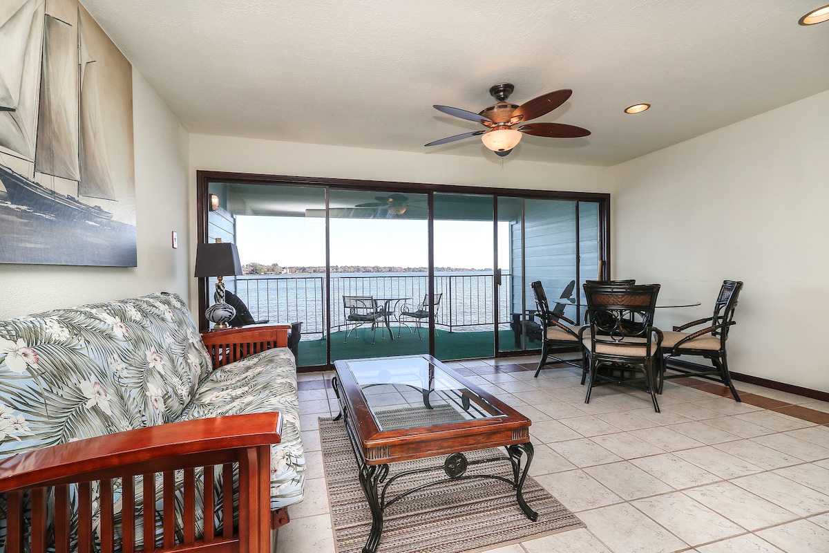 A living area features a tropical-themed sofa and a wooden coffee table, complemented by ceiling fans and recessed lighting. Large sliding glass doors provide a view of the lake, while a wooden dining set and additional seating area are positioned for comfort and relaxation.