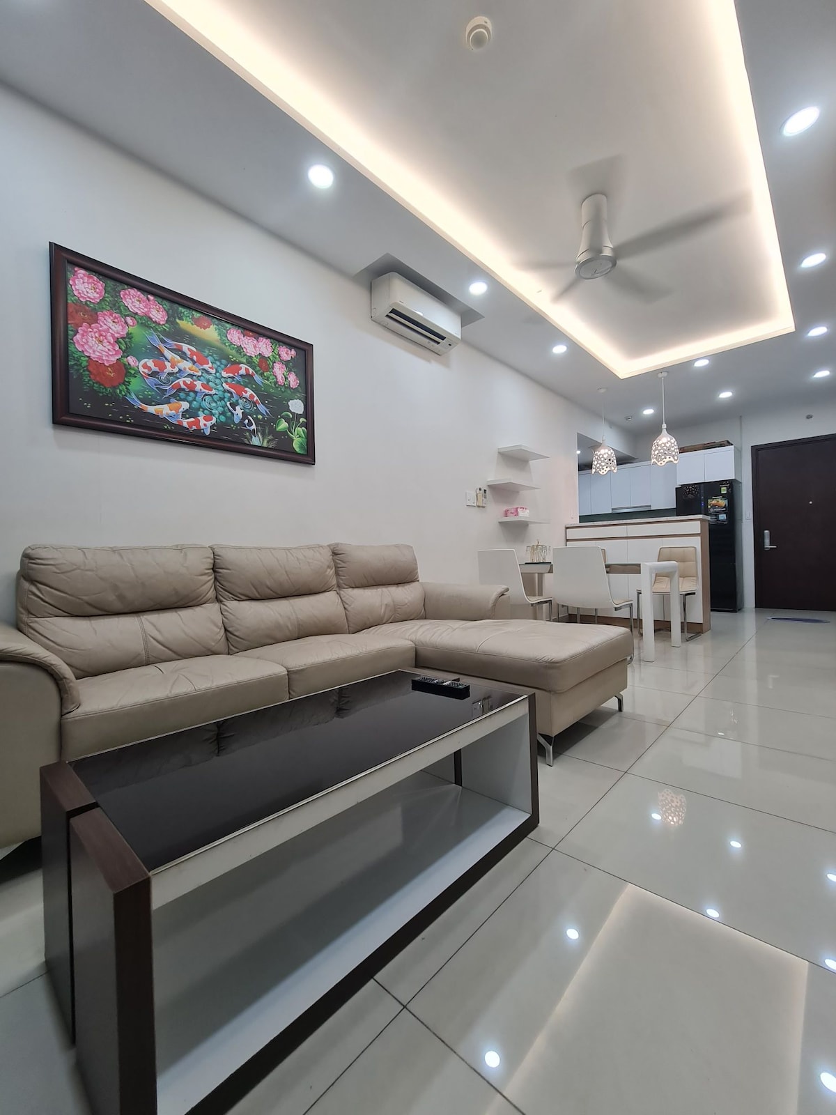 A modern living area features a light beige sectional sofa, arranged alongside a sleek coffee table. A colorful wall art piece adds visual interest. Bright lighting enhances the space, which transitions into a dining area with a table and chairs in the background.