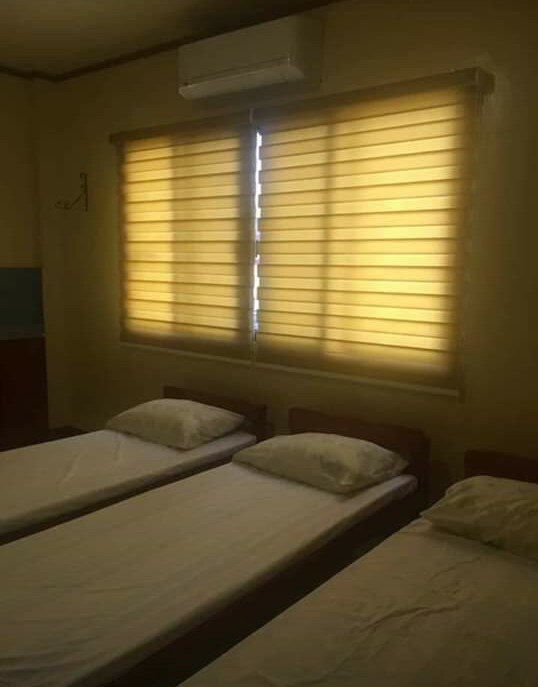 A comfortable sleeping area is furnished with three single beds, each topped with a pillow. Soft yellow blinds allow natural light to filter into the room, creating a warm ambiance. A wall-mounted air conditioning unit is visible above the beds.