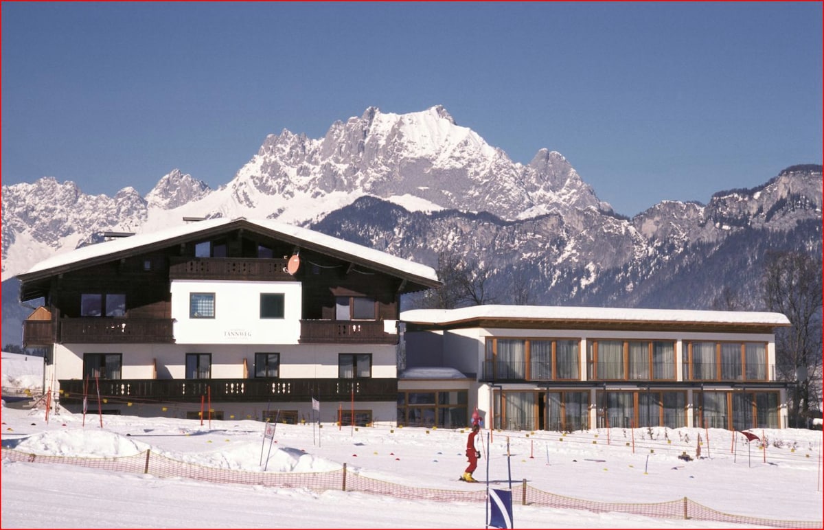 A chalet-style building is situated against a backdrop of snow-capped mountains. The structure combines traditional wooden accents with large glass windows, allowing natural light to brighten the interior. Surrounding areas are covered in snow, indicating a winter setting, with ski tracks visible in the foreground.