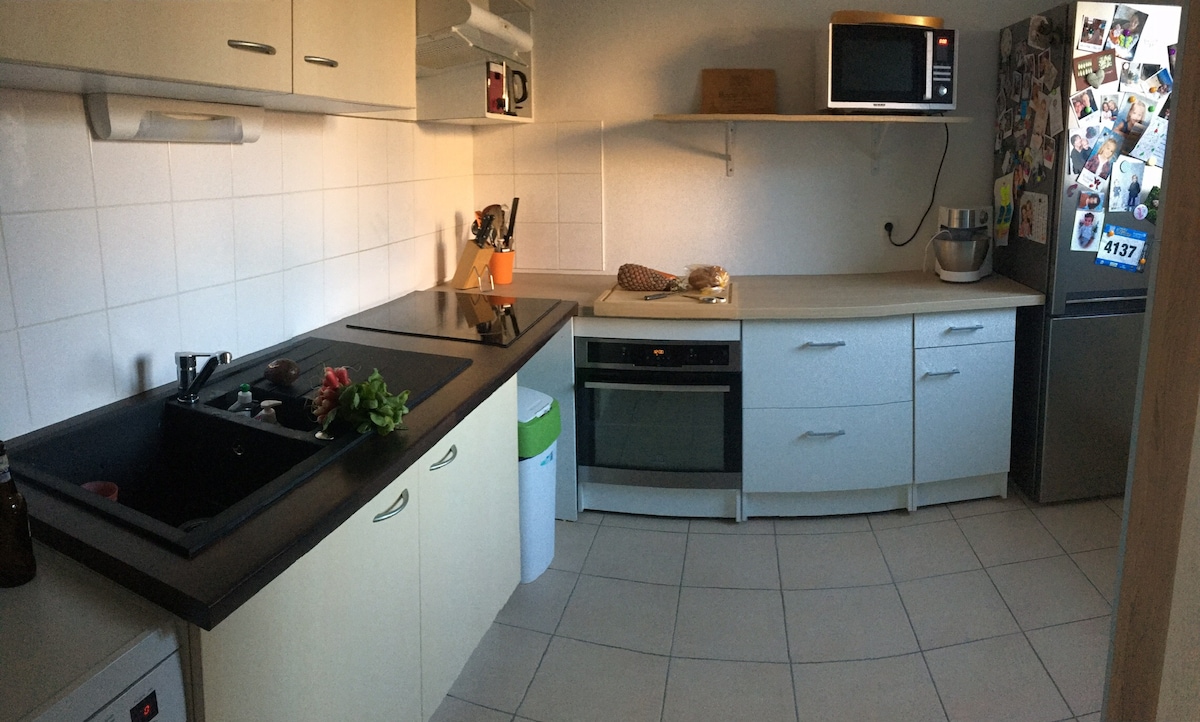 A well-equipped kitchen showcases modern appliances, including an oven, microwave, and refrigerator. Counter space features a sink, cooking utensils, and a small cutting board. White cabinets and tiled walls create a clean, organized appearance, with a waste bin positioned discreetly in the corner.