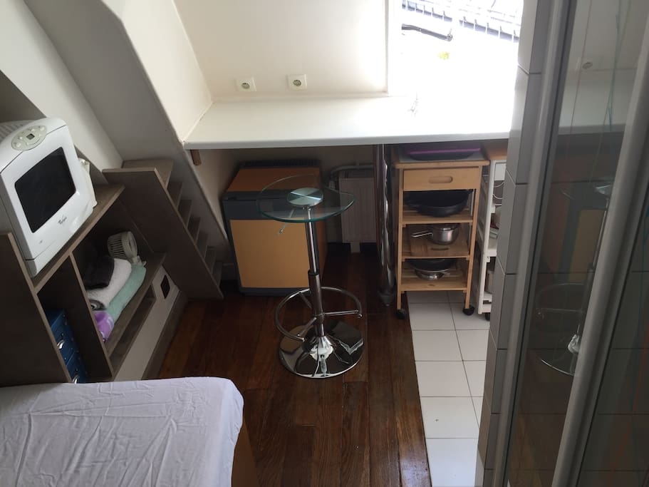 Overview of bed with white sheet, oak floors under bed and white tiles in kitchen and shower area