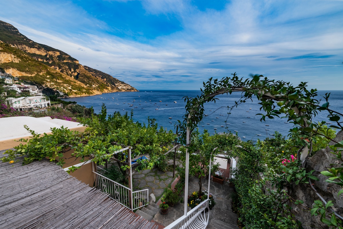A private terrace lined with lush greenery overlooks the clear blue waters of the Positano sea. The curved path leads down through a garden of vibrant plants, with the hillside and coastline creating a serene backdrop under a partly cloudy sky.