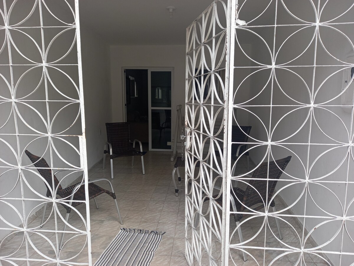 An entrance is framed by a decorative white metal gate, leading into a covered outdoor area with tiled flooring. Two wicker chairs are positioned on each side, creating a welcoming space. The interior is visible through glass doors, offering a glimpse of the living area.