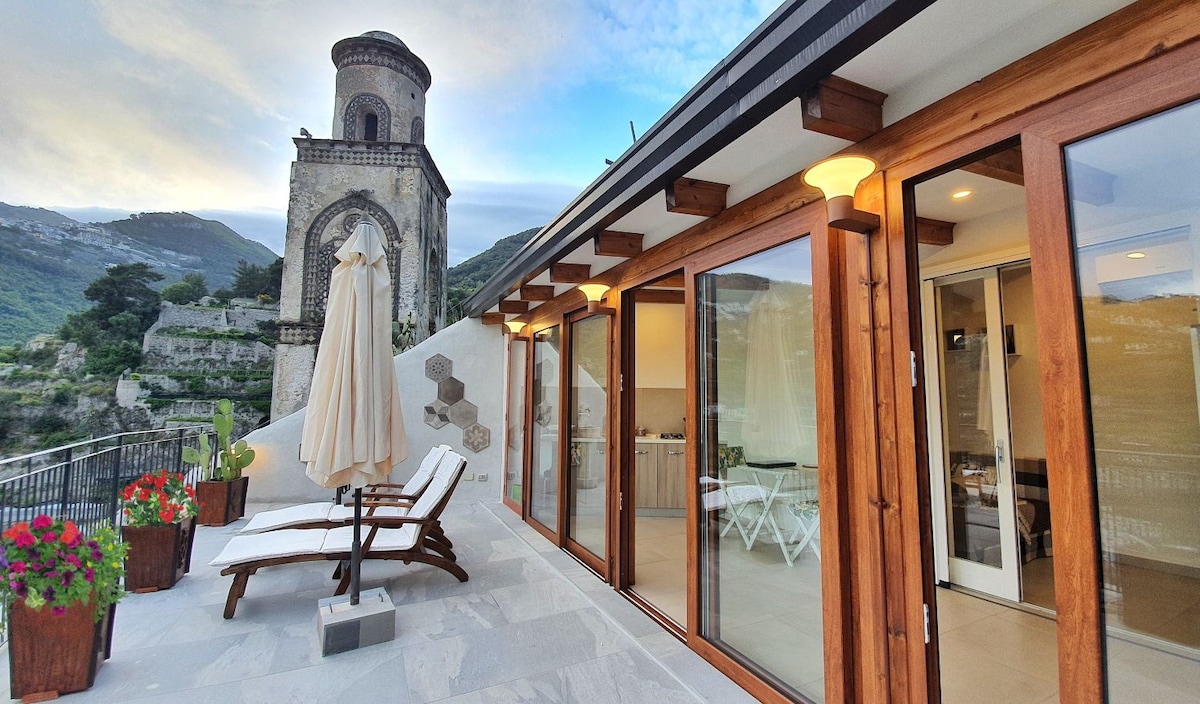 A spacious terrace is highlighted, featuring comfortable sun loungers and potted flowers. Large glass doors provide access to the interior, showcasing a modern kitchen area. In the background, an ancient bell tower stands against a mountainous landscape, adding character to the serene outdoor space.