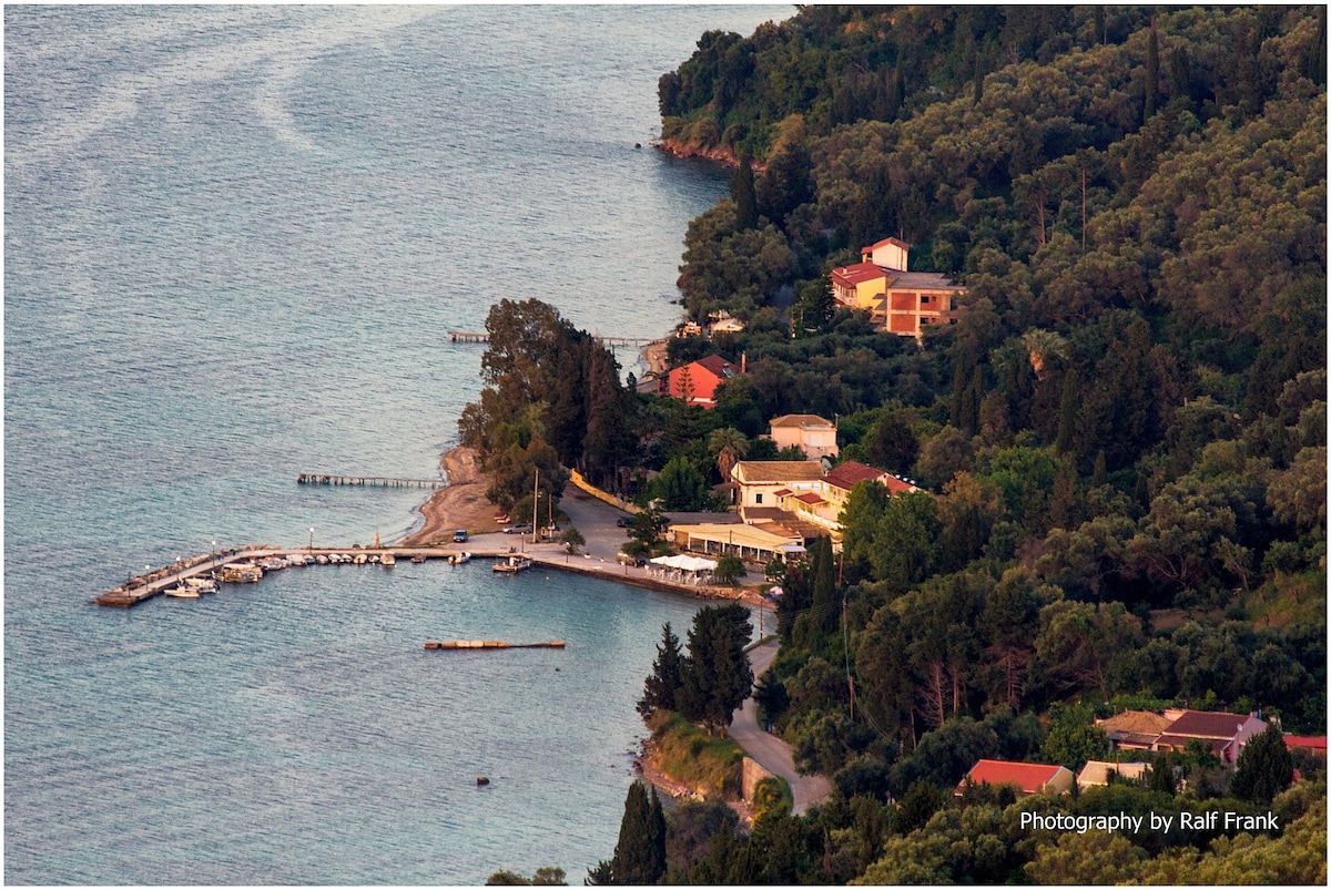 A coastal landscape is captured, featuring a serene shoreline with a small beach and a marina. Houses are nestled among lush greenery, while the calm sea reflects the surrounding natural beauty. A road curves along the coast, offering access to the tranquil environment.