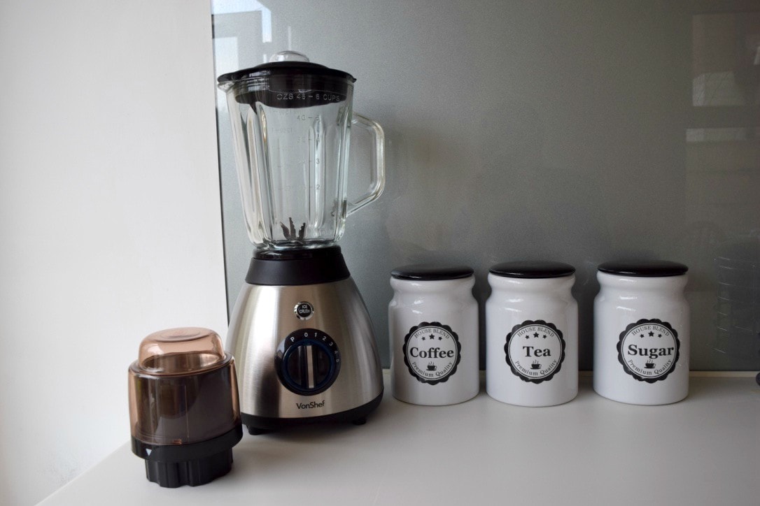 A sleek silver blender is positioned beside three white ceramic containers labeled 'Coffee', 'Tea', and 'Sugar'. The containers display simple black designs, adding a subtle contrast to the modern kitchen setting.