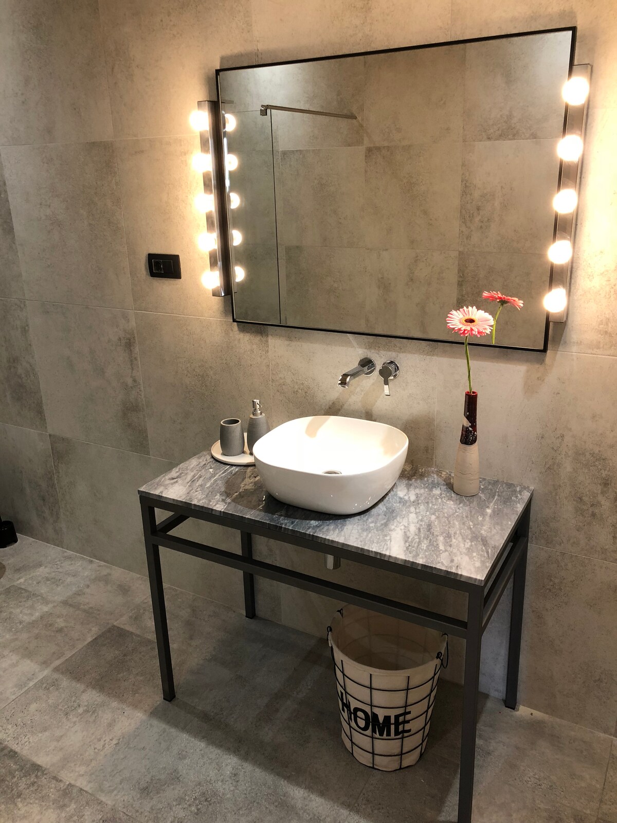 A modern bathroom setting features a sleek sink atop a marble countertop, complemented by a decorative vase with a single flower. Bright lights are positioned around the mirror, enhancing visibility. A stylish wastebasket is situated underneath, contributing to the overall tidy appearance.