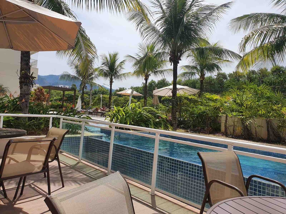 A serene outdoor space is presented, featuring a modern pool surrounded by lush greenery and palm trees. Sun loungers are placed beneath umbrellas, providing shaded areas for relaxation. The terrace includes several chairs arranged for comfort, complementing the tranquil setting.