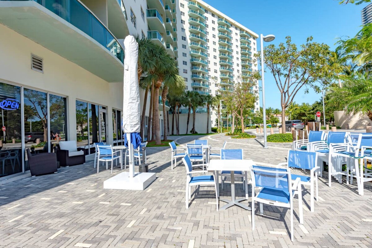 An outdoor patio area features several white dining tables and blue chairs arranged on a brick surface. A tall umbrella stand is positioned centrally, while palm trees provide shade in the background. The building's glass facade reflects the clear blue sky.