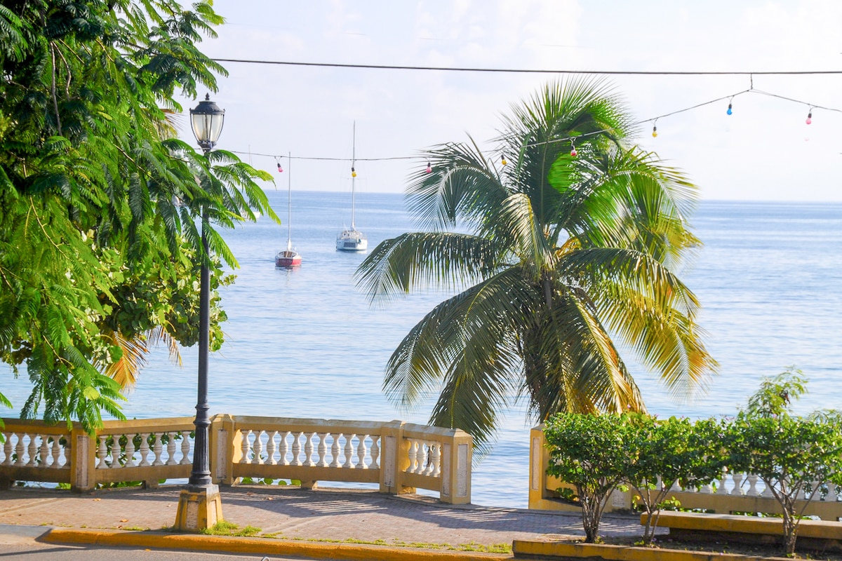 A serene waterfront scene features lush palm trees swaying gently, with a sparkling sea in the background. Two sailboats are anchored in the calm waters, surrounded by a picturesque coastal promenade adorned with decorative lights and vibrant greenery.