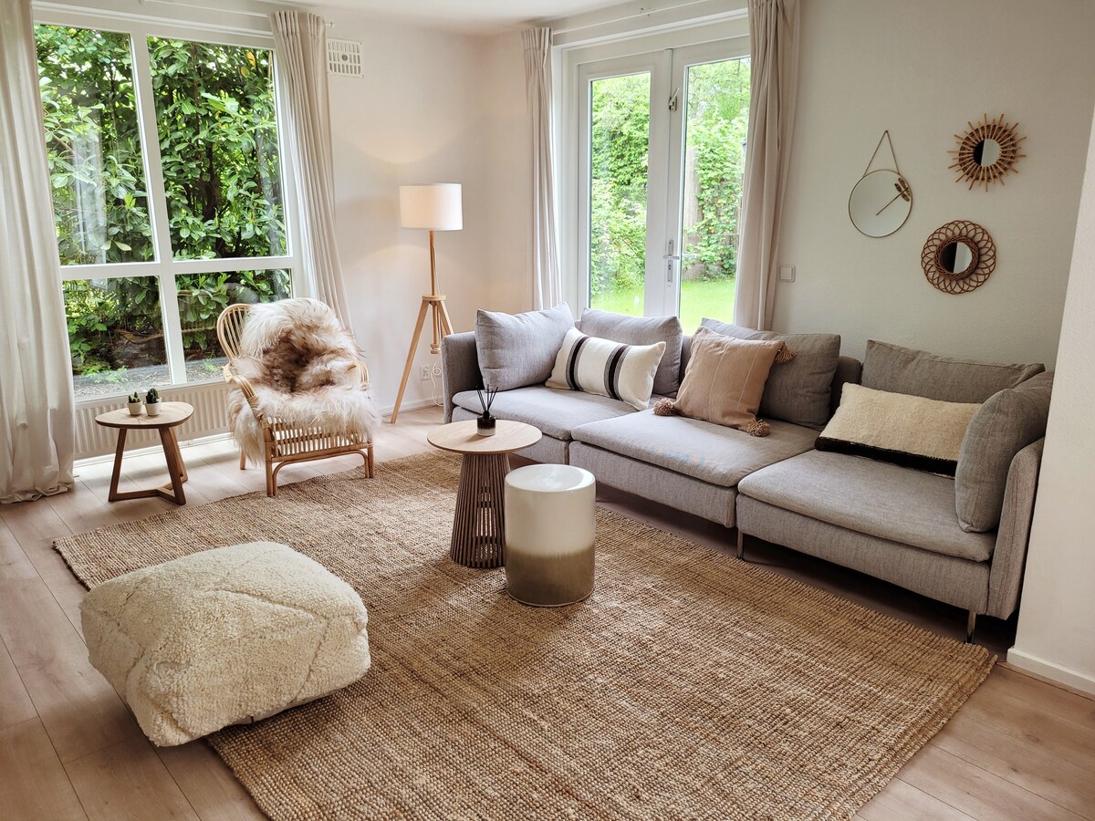 A well-lit living area features a comfortable gray sectional sofa with several decorative pillows. A cozy pouf and a round side table are situated on a natural fiber rug, while a stylish armchair provides additional seating. Large windows offer views of the greenery outside.
