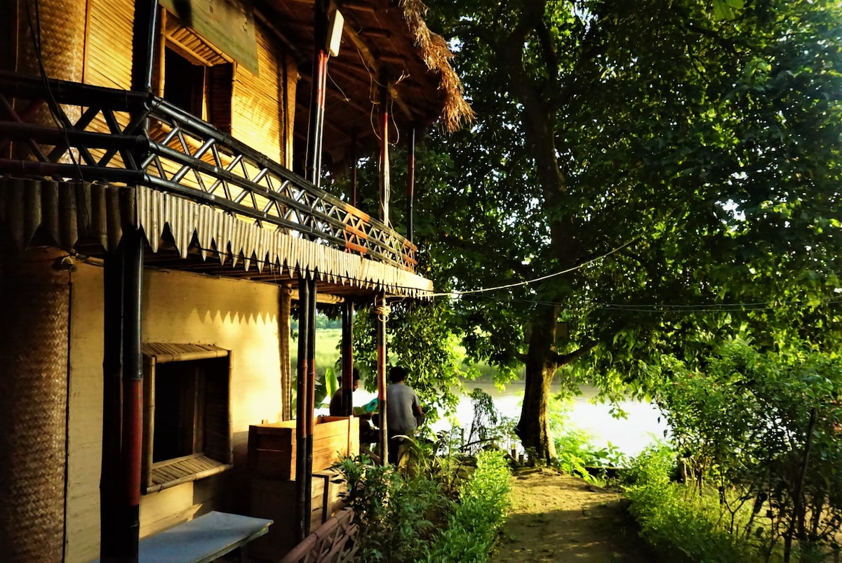 A bamboo lodge is depicted, showcasing a thatched roof and a spacious balcony adorned with natural wood railings. Lush greenery surrounds the structure, with a large tree casting shade. The riverbank is visible in the background, providing a serene natural setting.