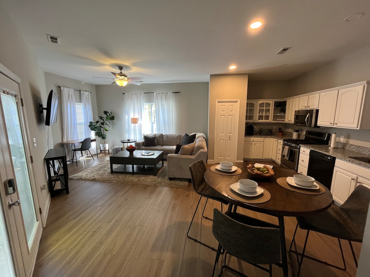 An open living area is showcased, featuring a large sectional couch facing a coffee table and a smart TV. A dining table is set for four, while a well-equipped kitchen is visible in the background. Natural light filters through sheer curtains.