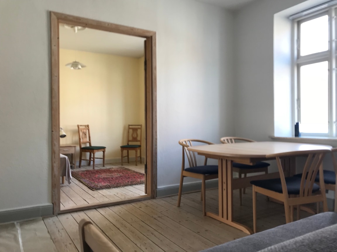A simple dining area features a round table surrounded by four wooden chairs. A doorway leads to a well-lit adjacent space with two additional chairs and a small rug on the floor. Natural light filters in through a nearby window.