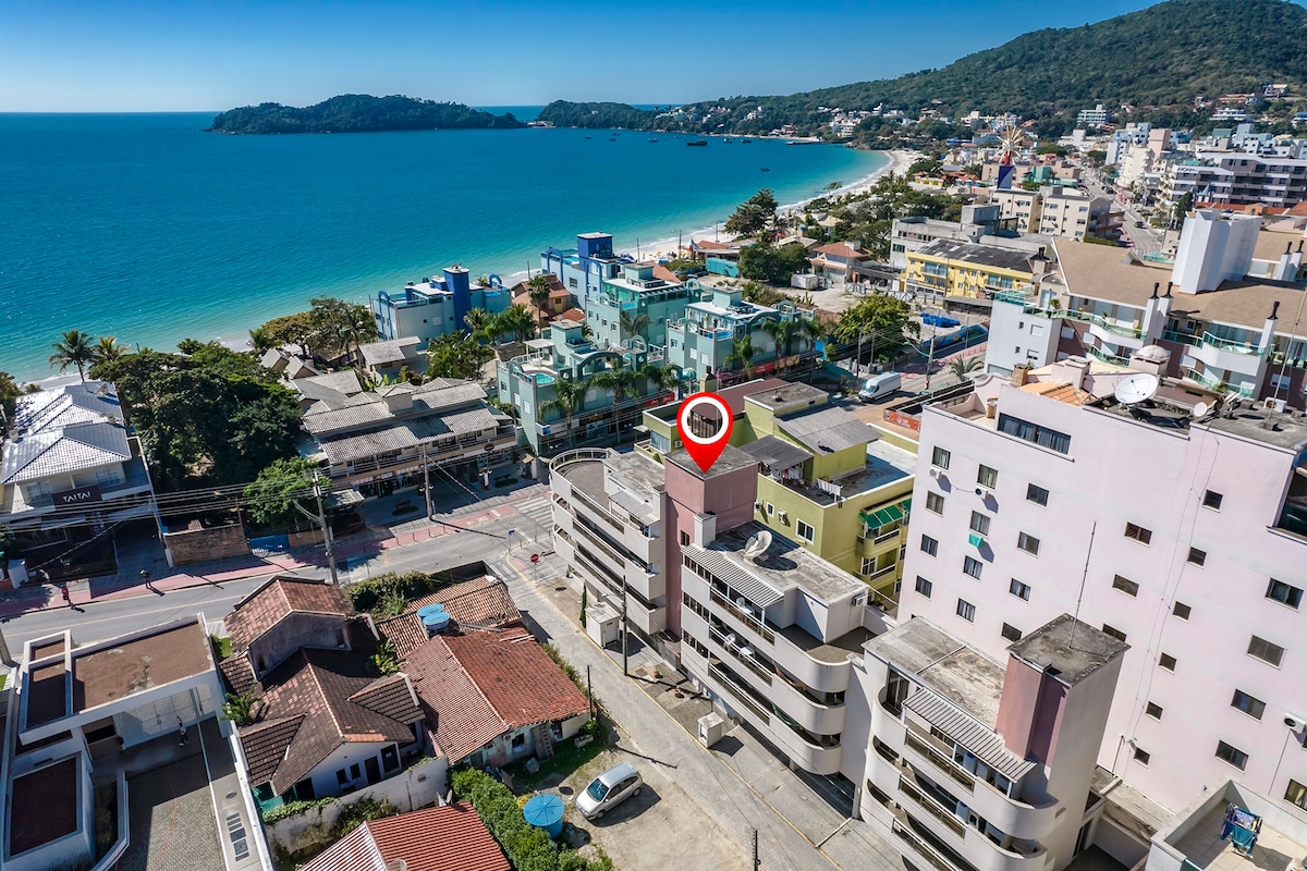 An aerial view captures the vibrant landscape of Bombinhas, showcasing the proximity to the coastline. The image features a mix of residential and commercial buildings, with the beach visible in the background and lush greenery lining the area.