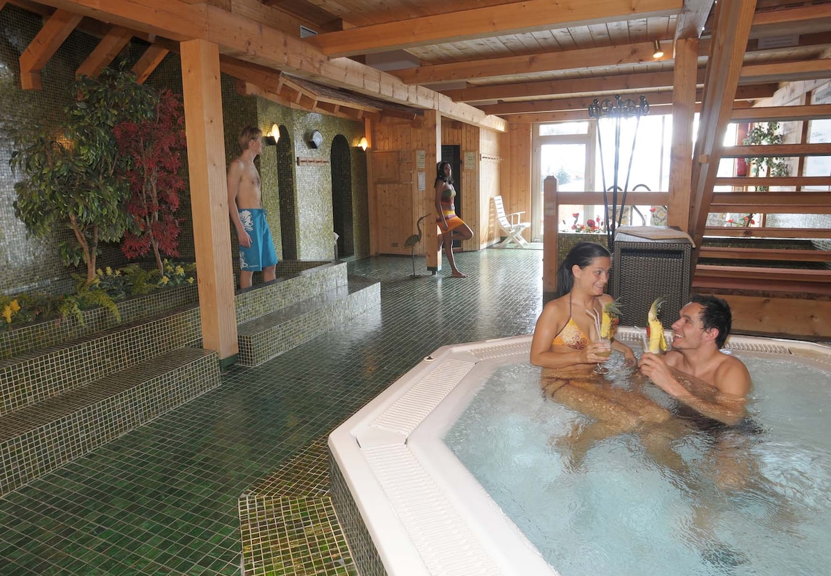 An inviting wellness area features a spacious indoor hot tub, surrounded by lush greenery and stone accents. Warm wooden beams reach above, enhancing the tranquil atmosphere. Soft lights create a serene ambiance, while a stairway leads to additional relaxation spaces in the backdrop.