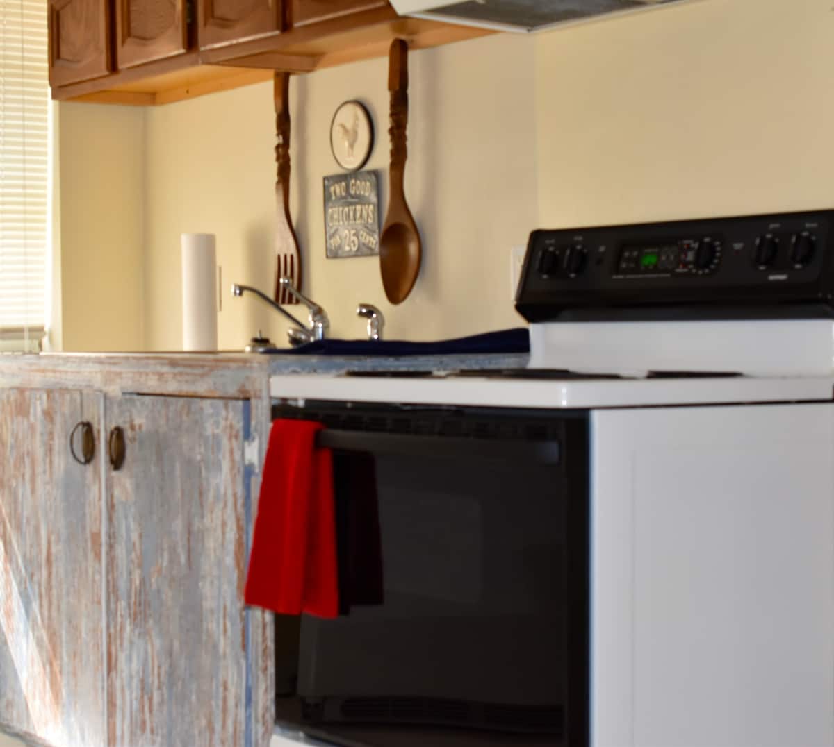 A kitchenette area is displayed featuring a combination of modern appliances and rustic cabinetry. A clean white stove and microwave are visible, alongside a decorative wooden sign and utensils hanging on the wall. A red towel is neatly hung on the cabinetry.