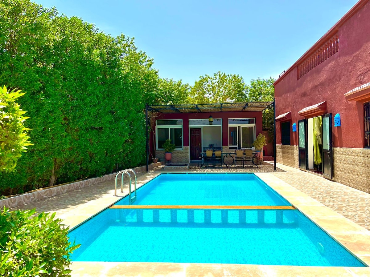 A refreshing swimming pool occupies the center of the outdoor area, surrounded by stone tiles. Green foliage provides privacy around the pool, while sliding glass doors lead into a cozy living space, which features seating and natural light from the windows.