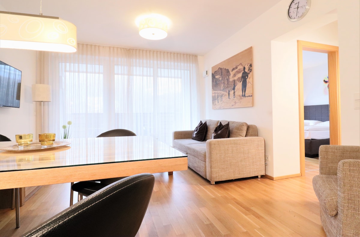 A bright living area features a glass dining table with black chairs and a cozy sofa. Large windows allow natural light to fill the space, complemented by soft curtains. A piece of artwork is displayed on the wall, adding character to the room.