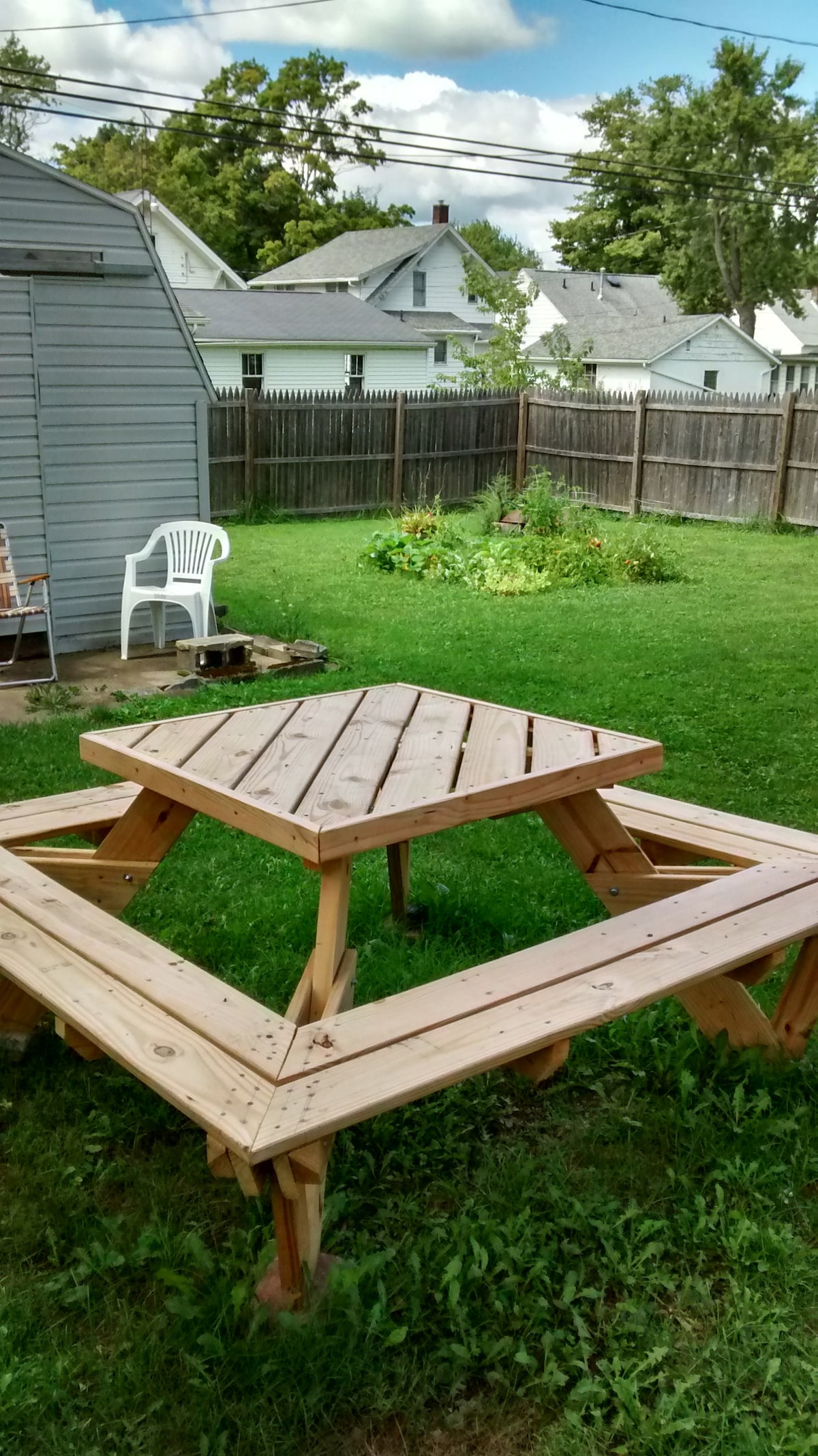 An outdoor area features a large wooden picnic table situated on grassy ground, surrounded by a fenced backyard. Sunlight casts gentle shadows on the table's flat surface, inviting potential for gatherings. A simple white chair and plant greenery are visible in the background.