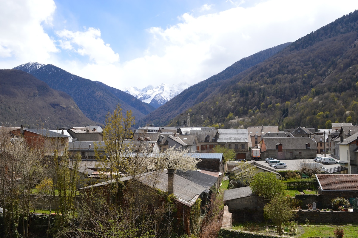 A scenic view of a mountain town is presented, surrounded by towering peaks, some capped with snow. Below, rooftops of local buildings are visible, interspersed with trees and gardens. The sky is partially cloudy, allowing sunlight to illuminate parts of the landscape.