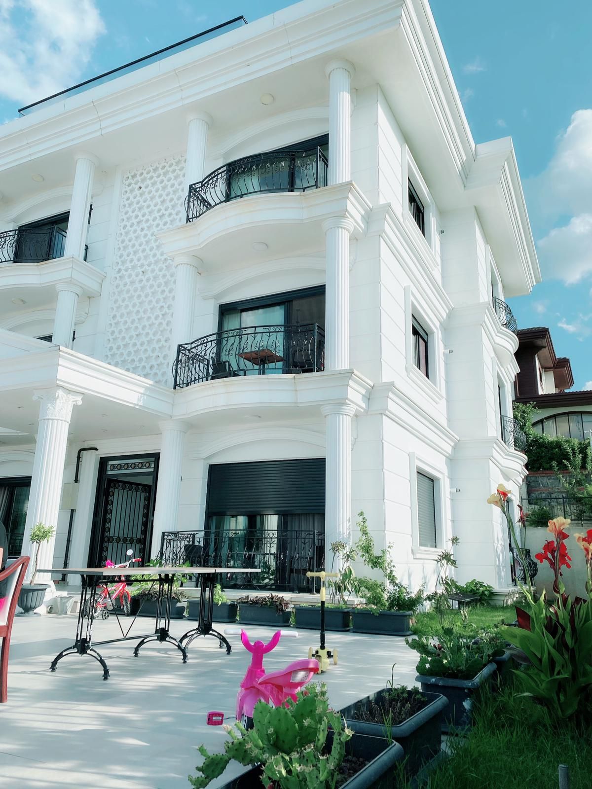 A modern, white multi-story villa features elegant balconies with wrought iron railings. Surrounding greenery and planted flowers enhance the outdoor space, highlighted by a patio area with tables and chairs. A playful pink flamingo adds a vibrant touch to the serene setting.