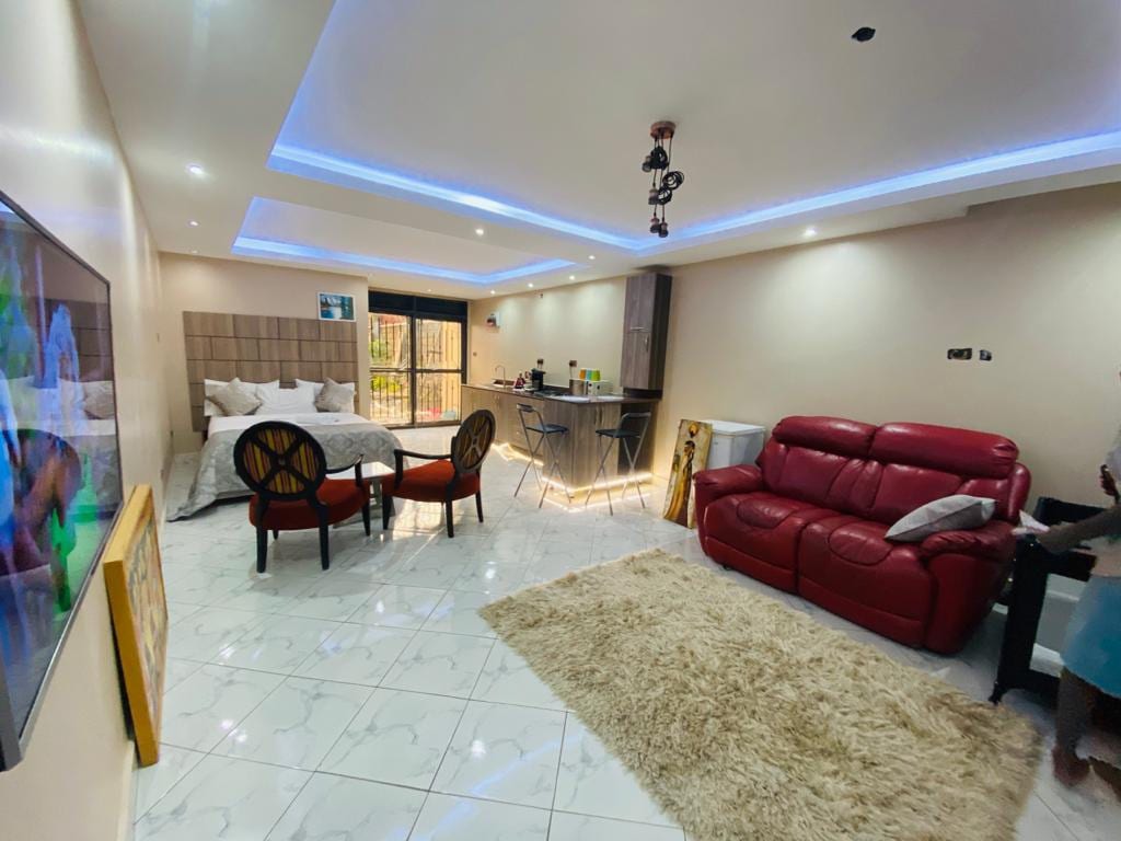 A contemporary studio suite features a spacious living area with a red sofa and stylish chairs. A modern kitchen is equipped with bar seating, while a comfortable bed area is visible in the background. Soft lighting accents the space, enhancing its inviting design.
