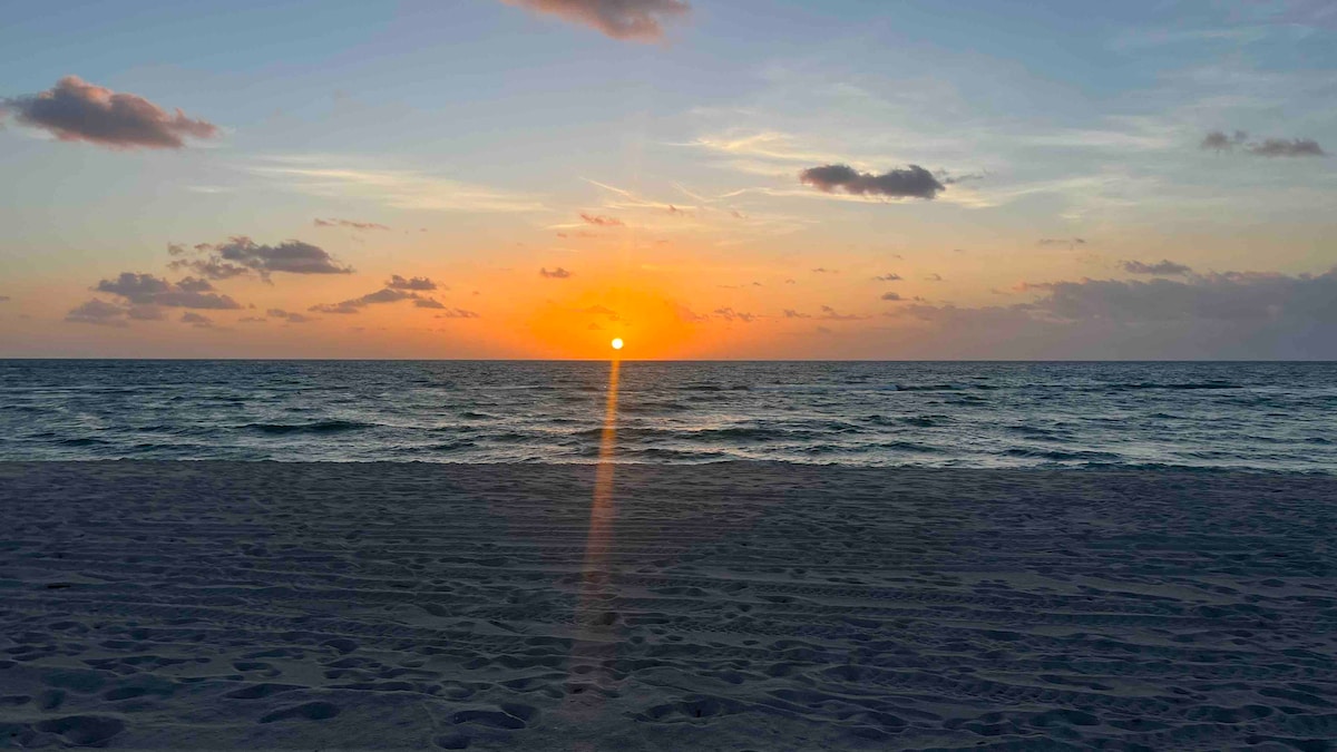 A calming sunset over the ocean is captured, with warm orange and yellow hues spreading across the sky. The sun rests just above the horizon, casting a reflective glow on the gentle waves. The sandy beach in the foreground leads to the water's edge.