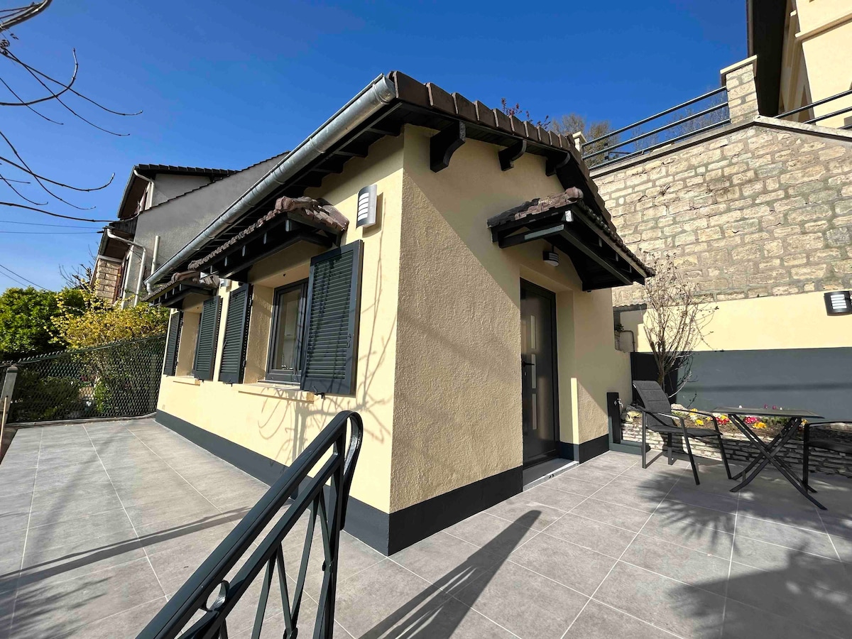 A modern, independent house is set against a clear blue sky. The exterior features a tiled roof, traditional shutters, and a spacious terrace adorned with outdoor seating. Grey tiles cover the terrace area, providing a clean and inviting entrance.