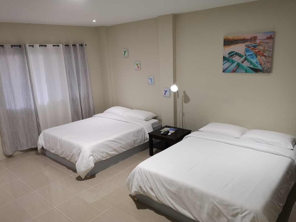 A cozy room is furnished with two spacious beds covered in white linens. Natural light filters through sheer curtains, illuminating the light-toned walls. A small bedside table holds refreshments, while colorful artwork of boats adorns one wall, adding a touch of character.